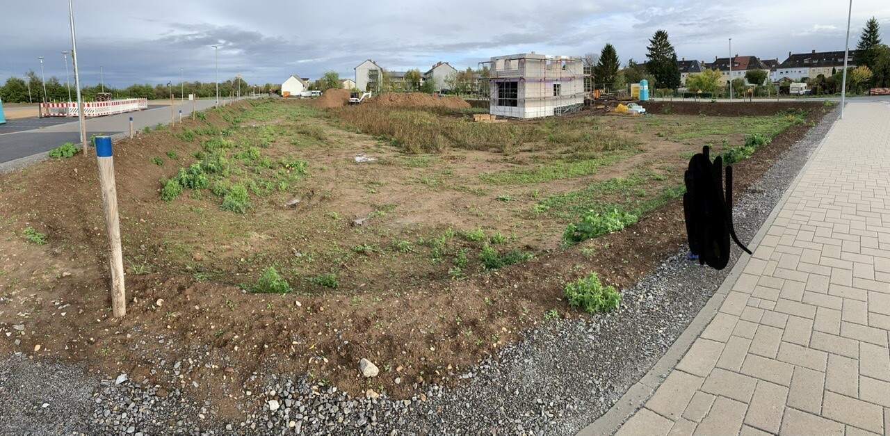 Großes Baugrundstück mit Erde, Grünflächen und Baukonstruktion im Hintergrund, rechter Gehweg.