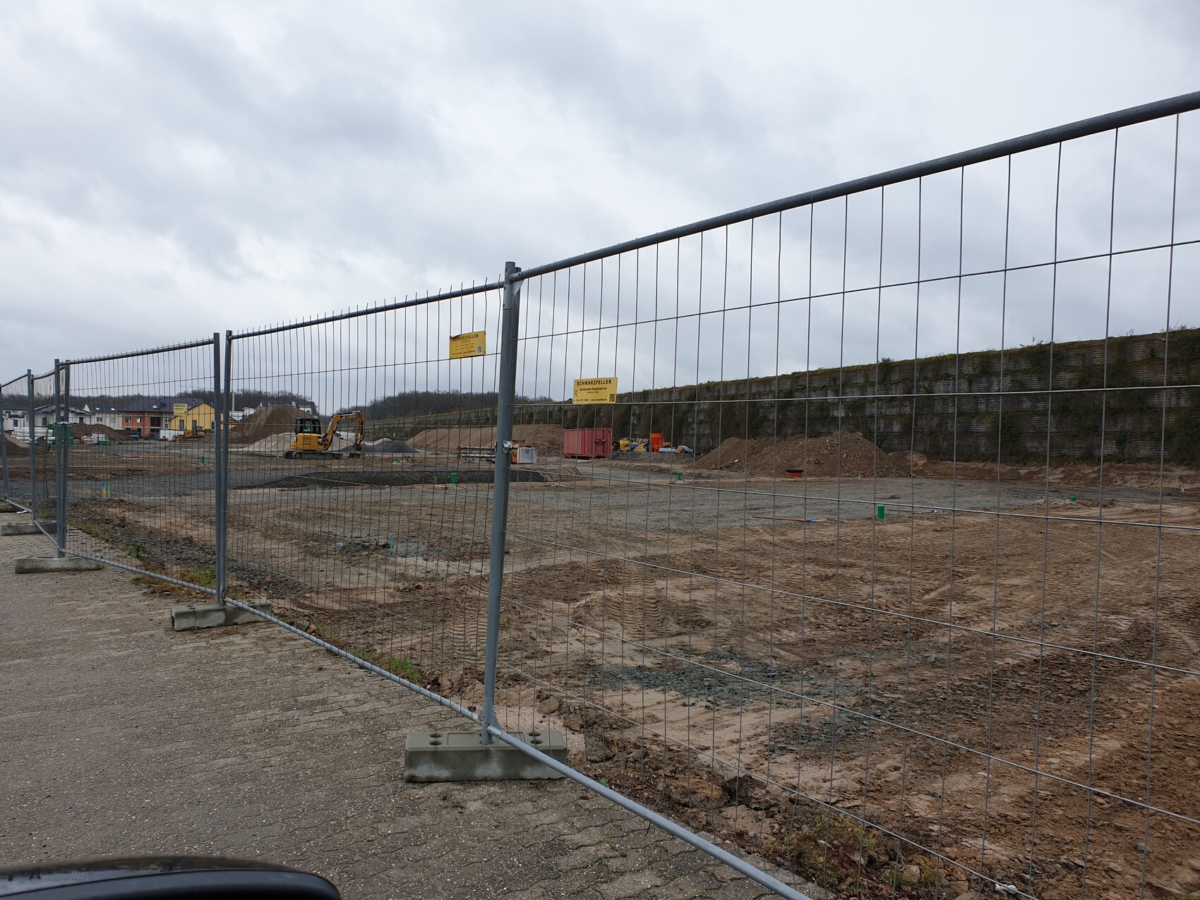 Baustelle hinter einem Metallgitter-Zaun; Bagger, Sandhaufen und bewölkter Himmel.