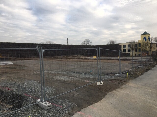 Leere Baustelle hinter Metallzaun, Erdarbeiten, Weg und Gebäude im Hintergrund.