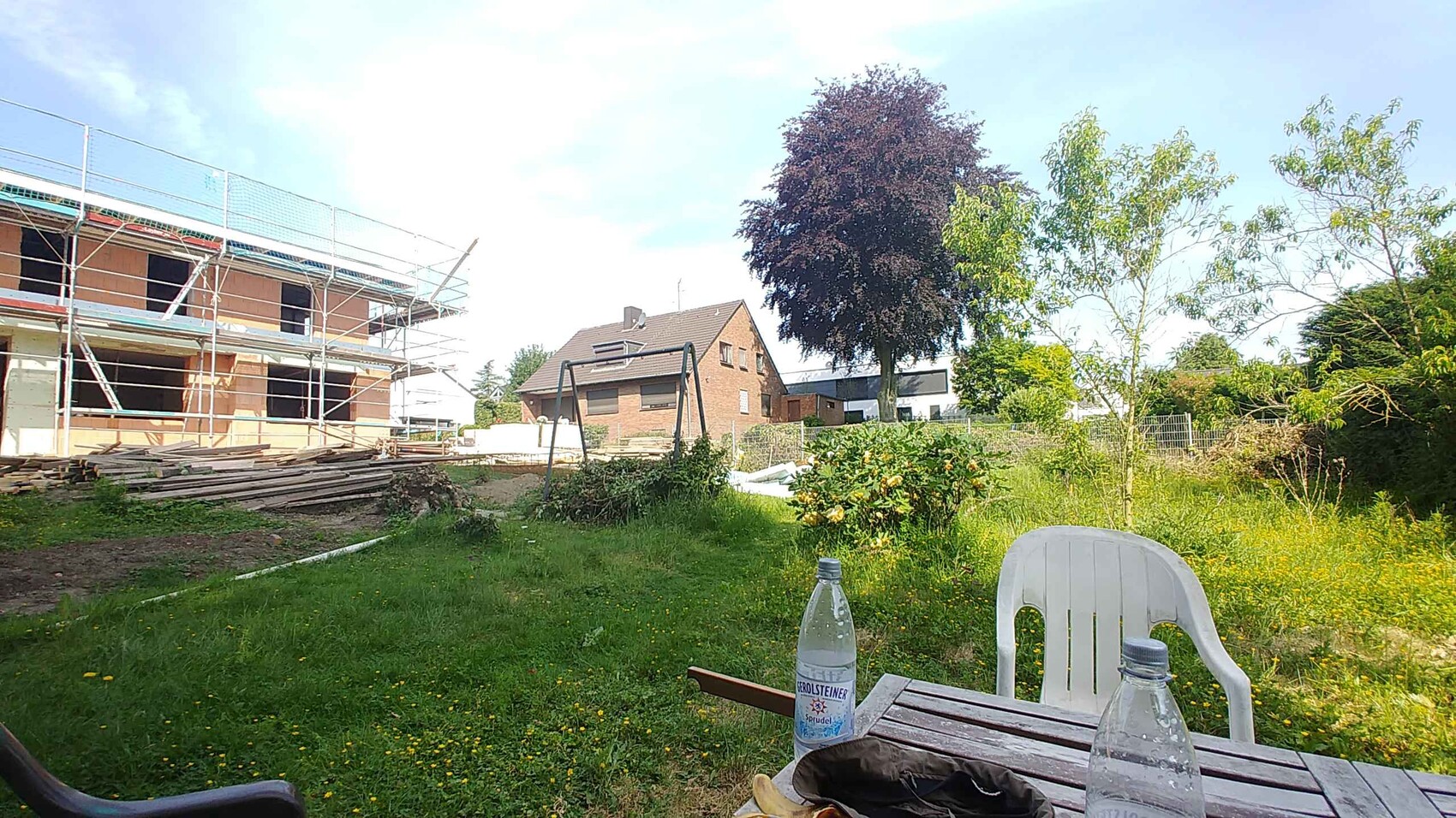 Baustelle mit Gerüst um rohbauendes Haus, Holzstapel, Garten und Bäume im Umfeld