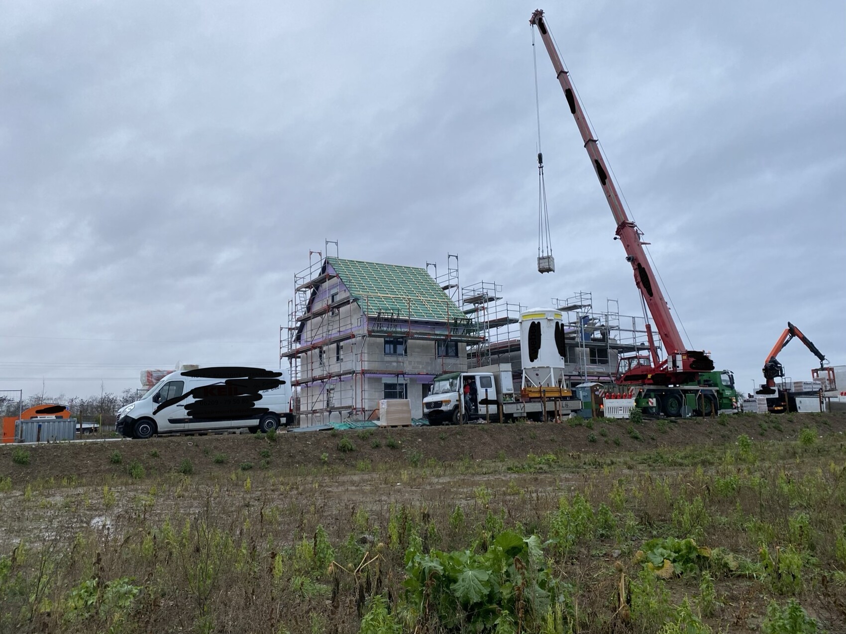 Baustelle: Einfamilienhaus-Rohbau mit Gerüst, Kran hebt Bauteil, Bauwagen