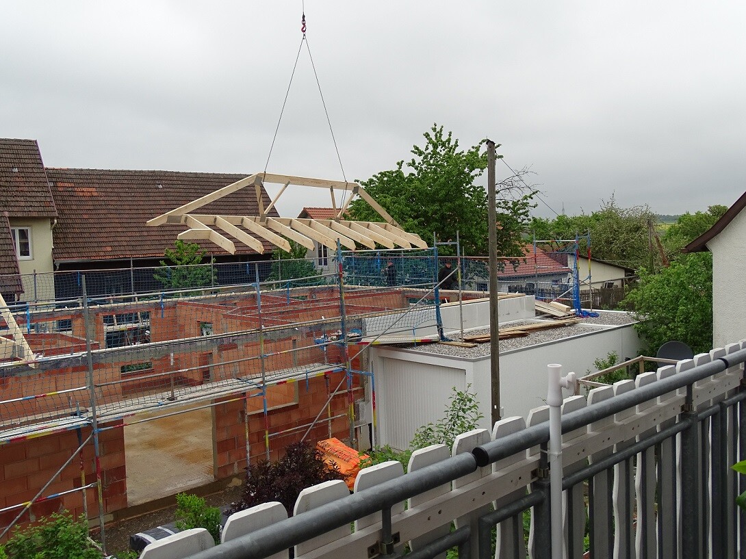 Baustelle mit Gerüsten; ein Holz-Dachstuhl wird per Kran auf einen Rohbau gesetzt.