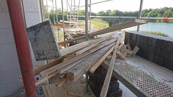 Baustelle mit Holzlatten, Betonblöcken und Gerüst; Wasser im Hintergrund.
