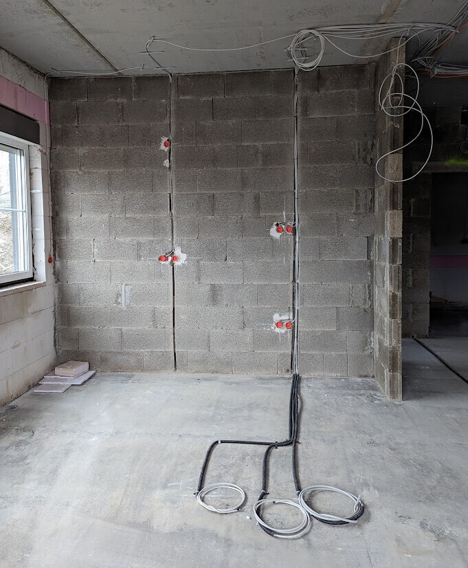 Rohbaustelle: unverputzte Ziegelwand, freiliegende Leitungen, rote Steckdosen, Kabel auf dem Boden.