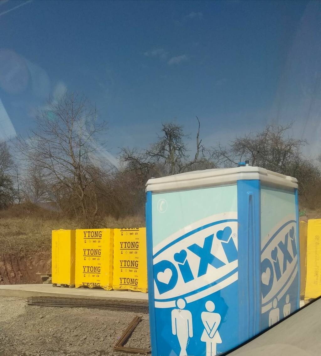 Baustellenszene: blaue DIXI-Toilette im Vordergrund, gelbe YTONG-Boxen links, kahle Bäume.