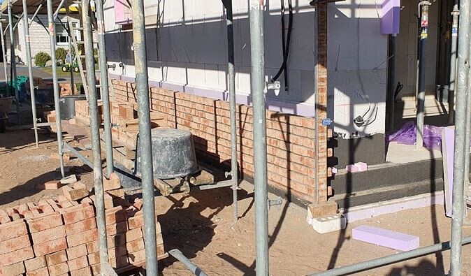 Baustelle mit Gerüst, unverputzte Ziegelwand, gestapelte Ziegel, Eimer und Erde.