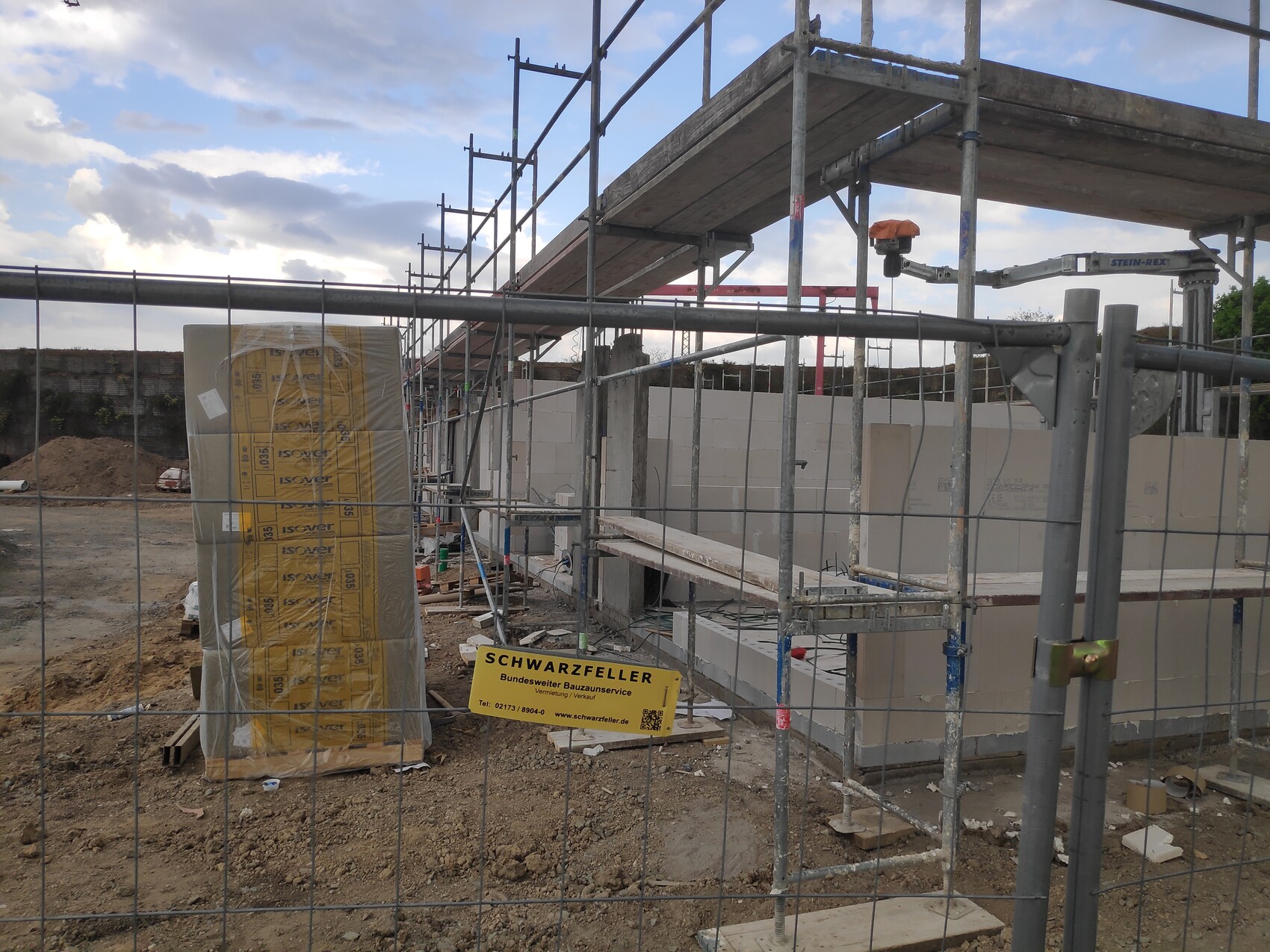 Baustelle mit Gerüst, unfertigen Betonwänden, gelb verpackte Isolierplatten auf Palette und Bauzaun.