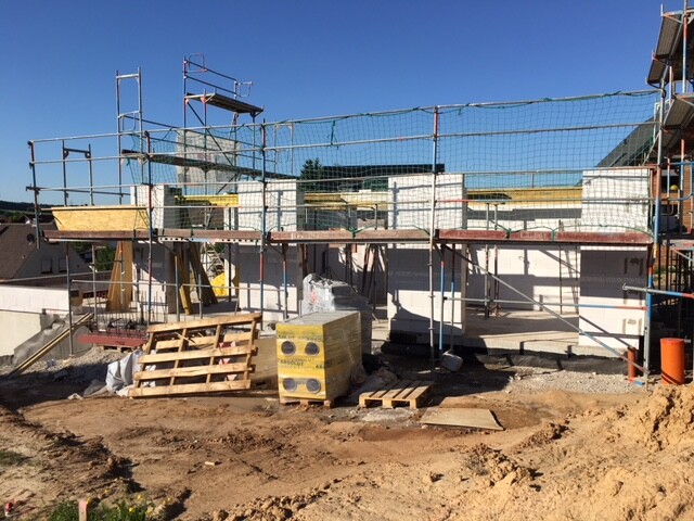 Baustelle mit Gerüst um ein unfertiges Gebäude, Holzpaletten und Baumaterialien im Freien.