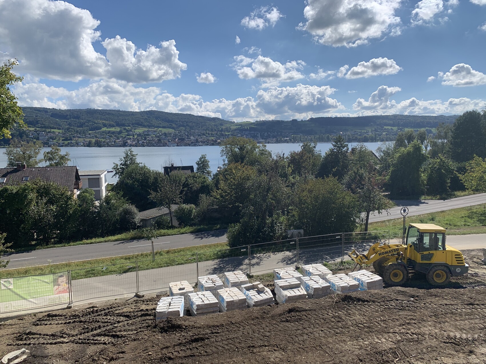 Baustelle am Hang: gelber Frontlader neben gestapelten Paletten, Blick auf See und Hügel.