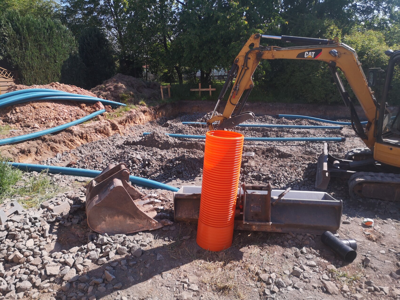 Gelber Bagger in Baugrube, orange Rohre und blaue Leitungsrohre liegen auf Kies.