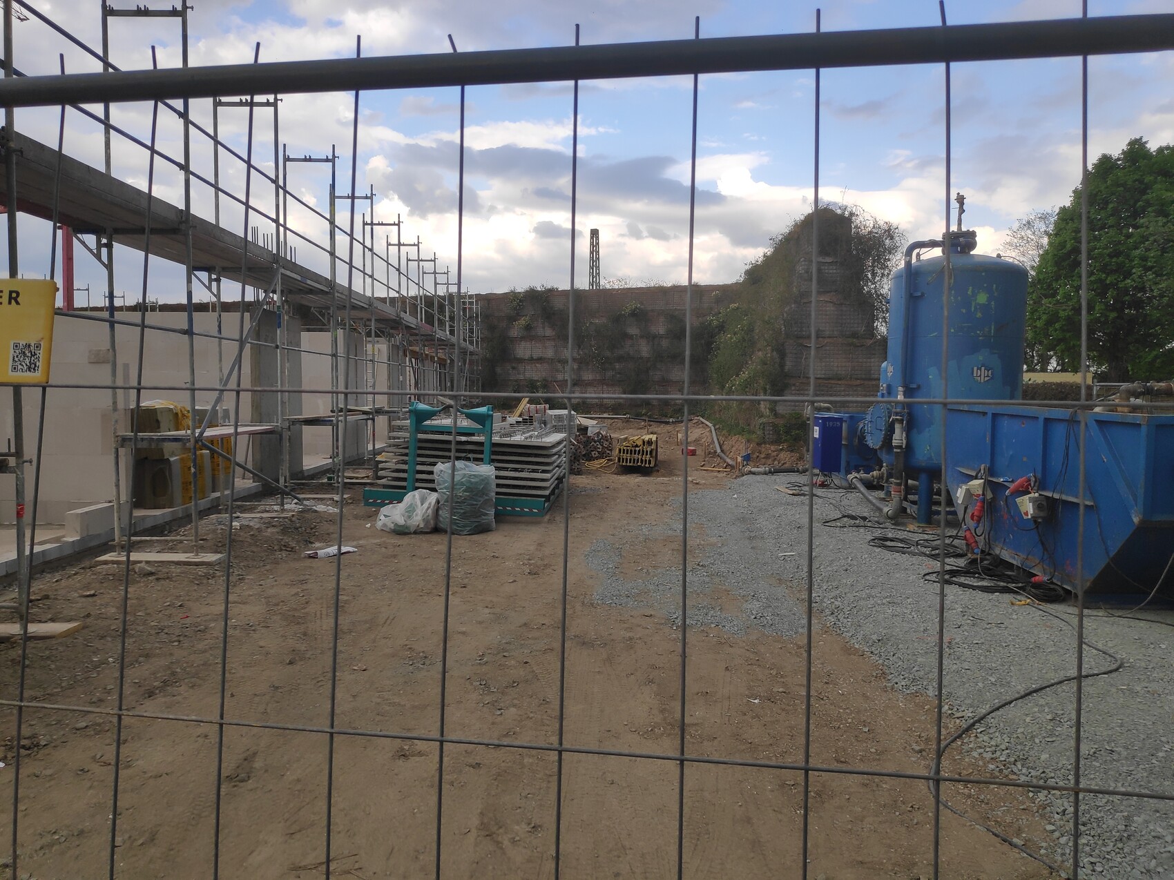 Baustelle hinter Zaun: Gerüst, Bauholz und Materialien; rechts blauer Tank, Wolken am Himmel