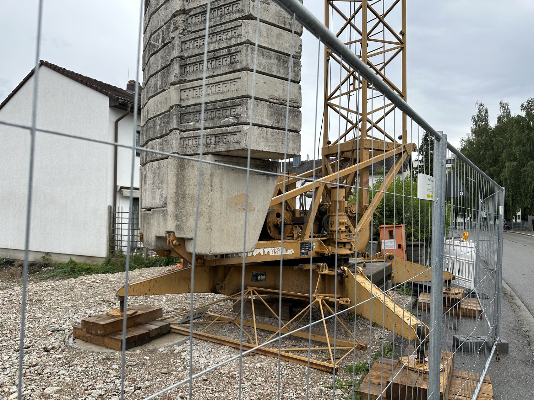 Große Pfahlbohrmaschine auf einer Baustelle hinter einem Metallzaun, Betonblöcke gestapelt.