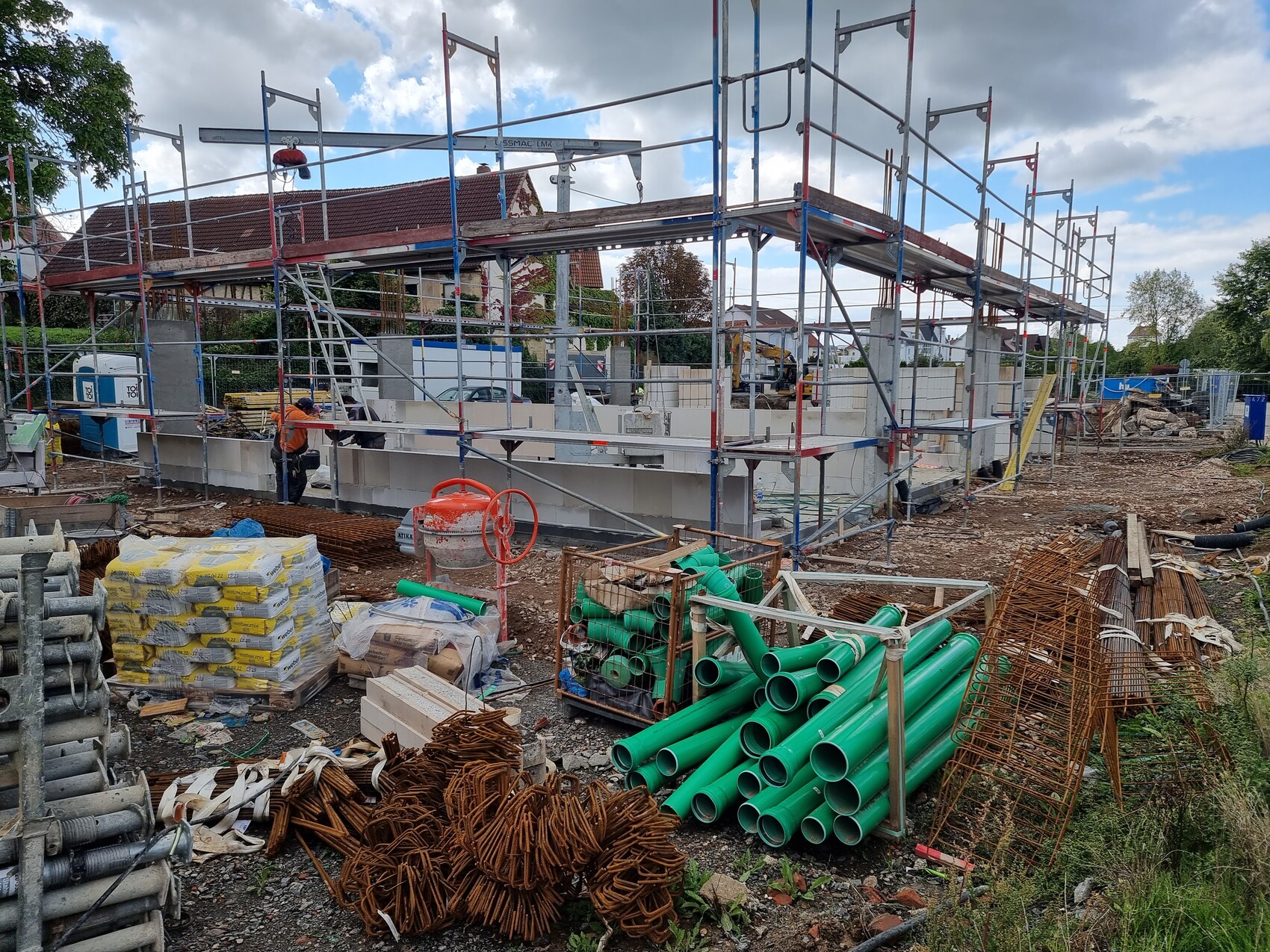Baustelle mit Gerüst, Arbeiter in Orange, grüne Rohre und Baumaterialien.