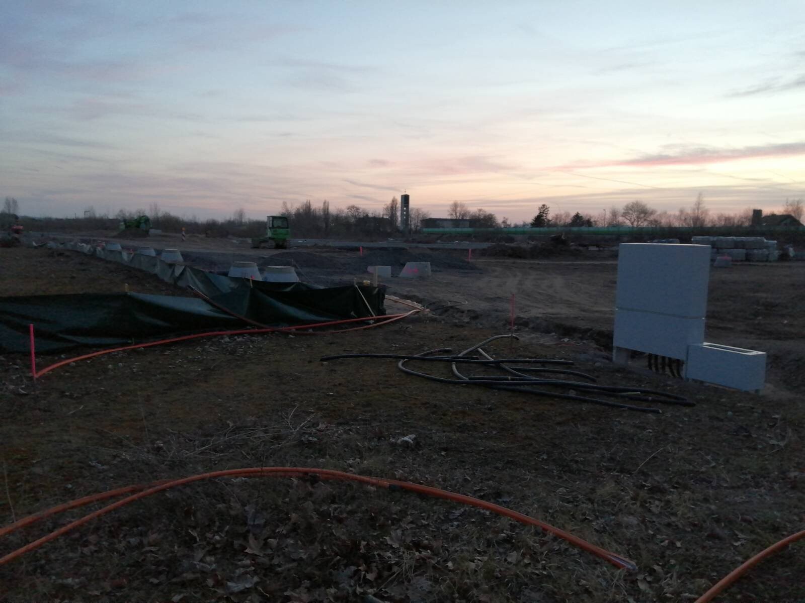 Baustelle im Feld bei Sonnenuntergang; Rohre, Kabel, Planen und Betonblöcke sichtbar.