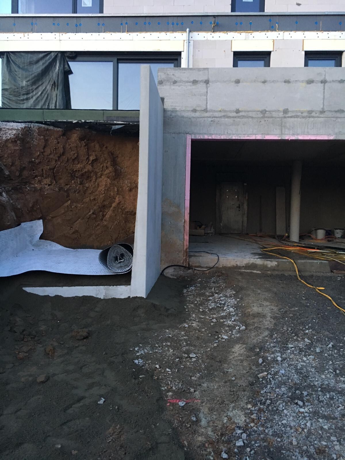 Baustelle mit offener Baugrube, Betonwänden, Kabeln und Geröll vor Gebäude.