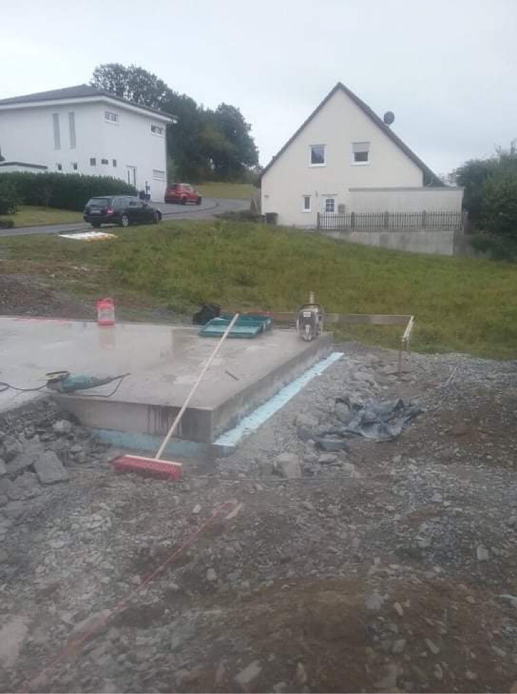 Baustelle mit Fundamentplatte, Schutt und Werkzeug; Wohnhäuser im Hintergrund.