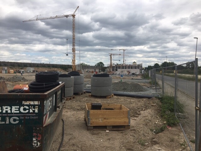Große Baustelle mit zwei Kränen, Reifenstapeln, Rohren und Holzkisten; Bauzaun rechts.
