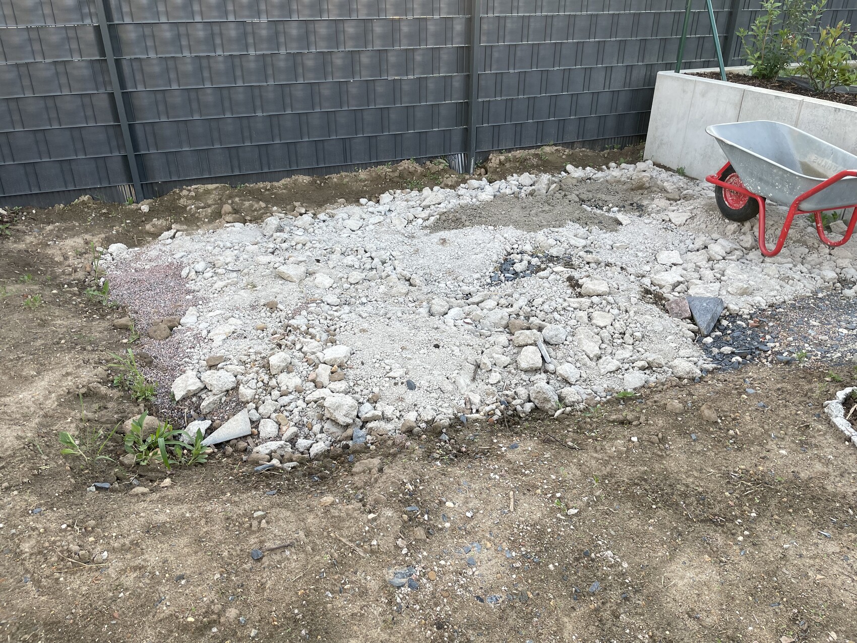 Baustelle im Garten: Kies, Geröll und Erdaushub, rote Schubkarre neben dunklem Zaun.