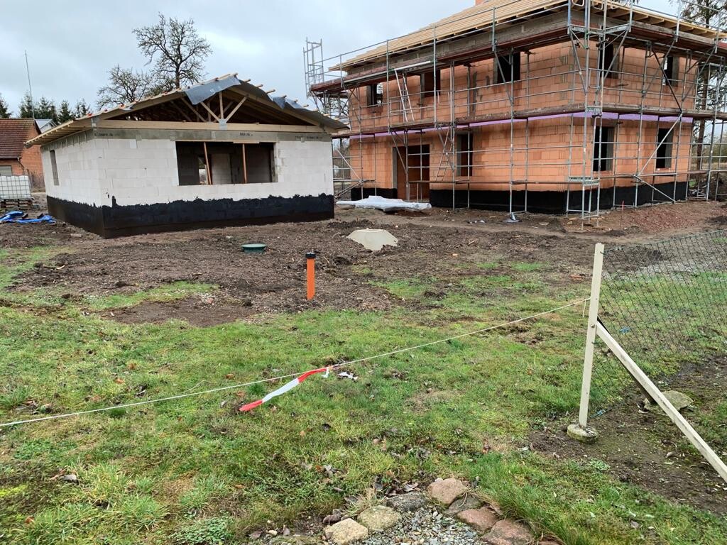 Baustelle: kleiner weißer Blockbau mit schwarzer Sockelabdichtung neben Rohbau mit Gerüst.