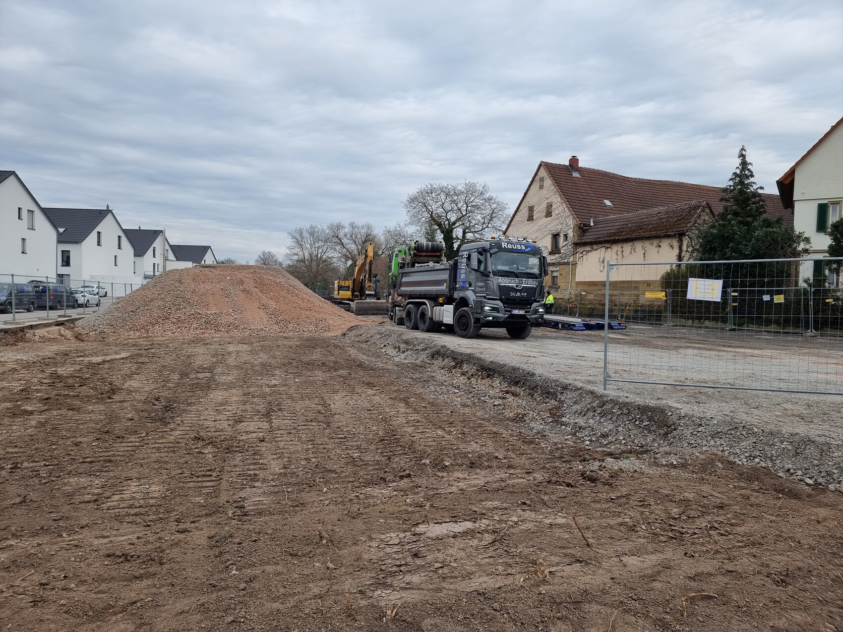 Großer Kieshaufen auf einer Baustelle mit Lastwagen und Baumaschinen vor Wohnhäusern.