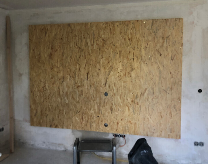 Unbearbeiteter Raum mit OSB-Platte an Wand, Leiter davor und Müllbeutel auf dem Boden.