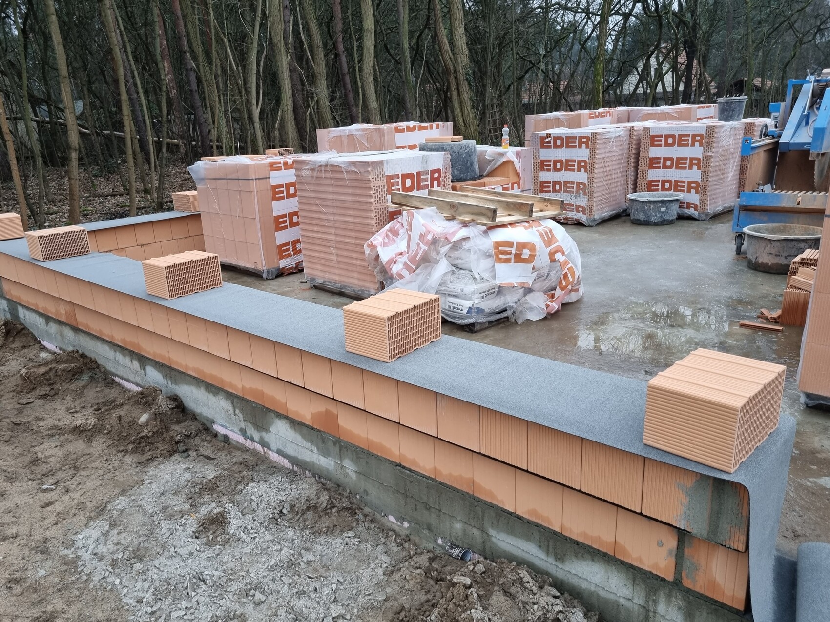 Baustelle mit gestapelten orangefarbenen Ziegeln, Paletten, Mörtelbehälter und Wald im Hintergrund.