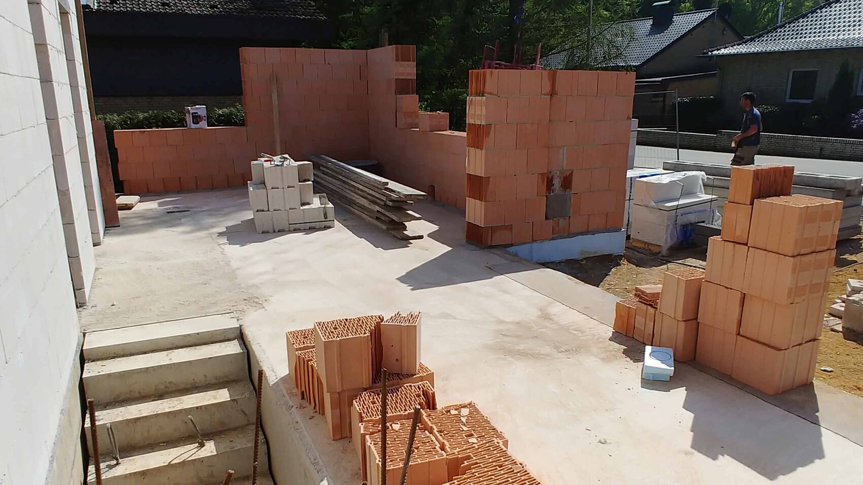 Außenbaustelle mit gestapelten orangefarbenen Ziegelsteinen, Holzplanken und Bauarbeiter rechts.