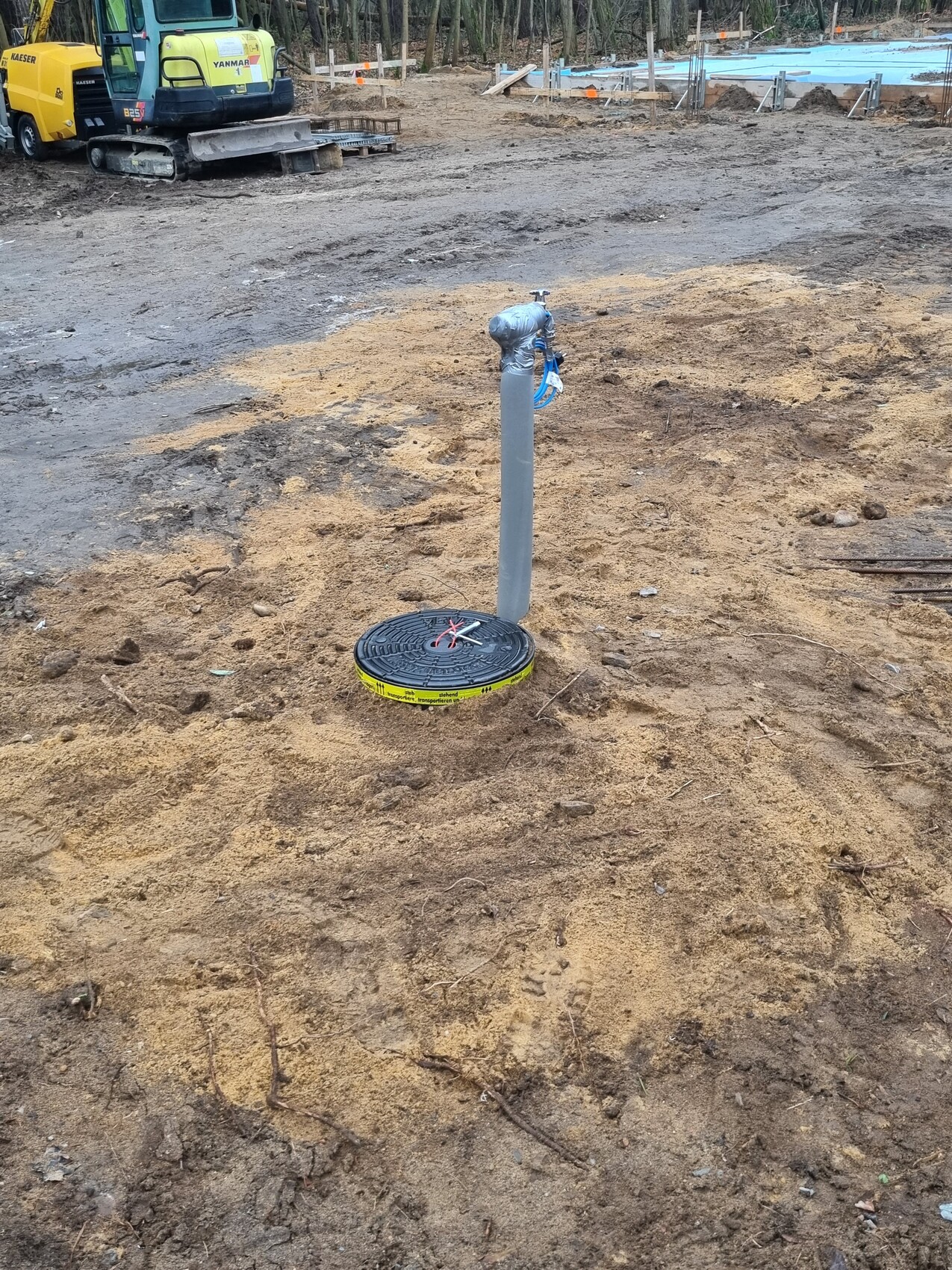 Baustelle: vertikales Wasserrohr aus sandigem Boden; gelb-schwarzer Deckel, Baugeräte im Hintergrund.