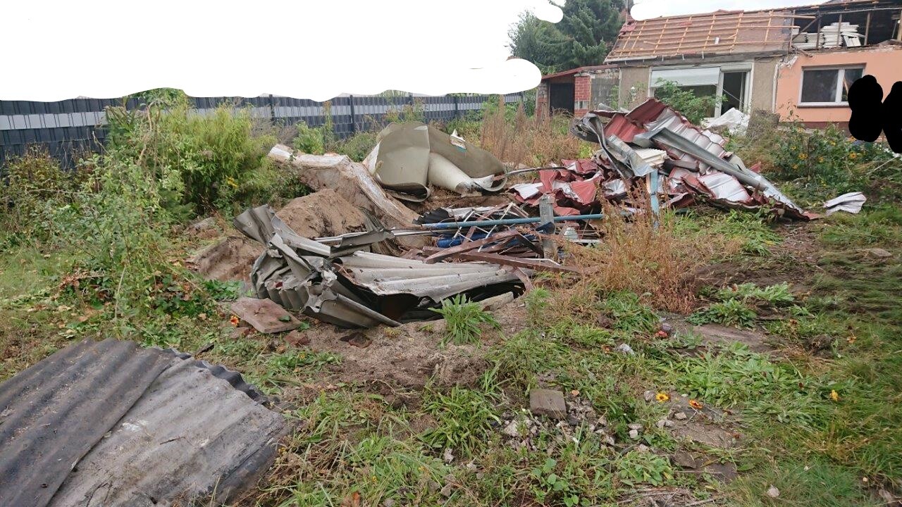 Garten mit Bauschutt und zerknülltem Dachmaterial, Unkraut, Haus im Hintergrund.