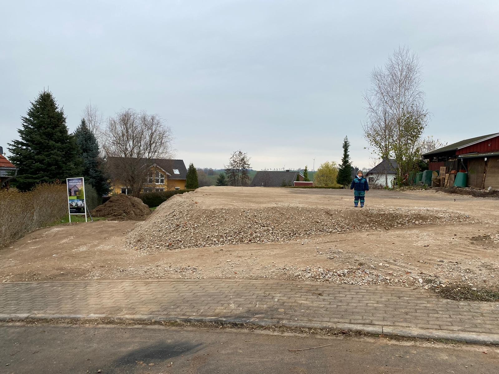 Kleiner Junge in blauem Mantel steht auf unbefestigtem Bauplatz mit Erdhügeln, Häuser im Hintergrund.