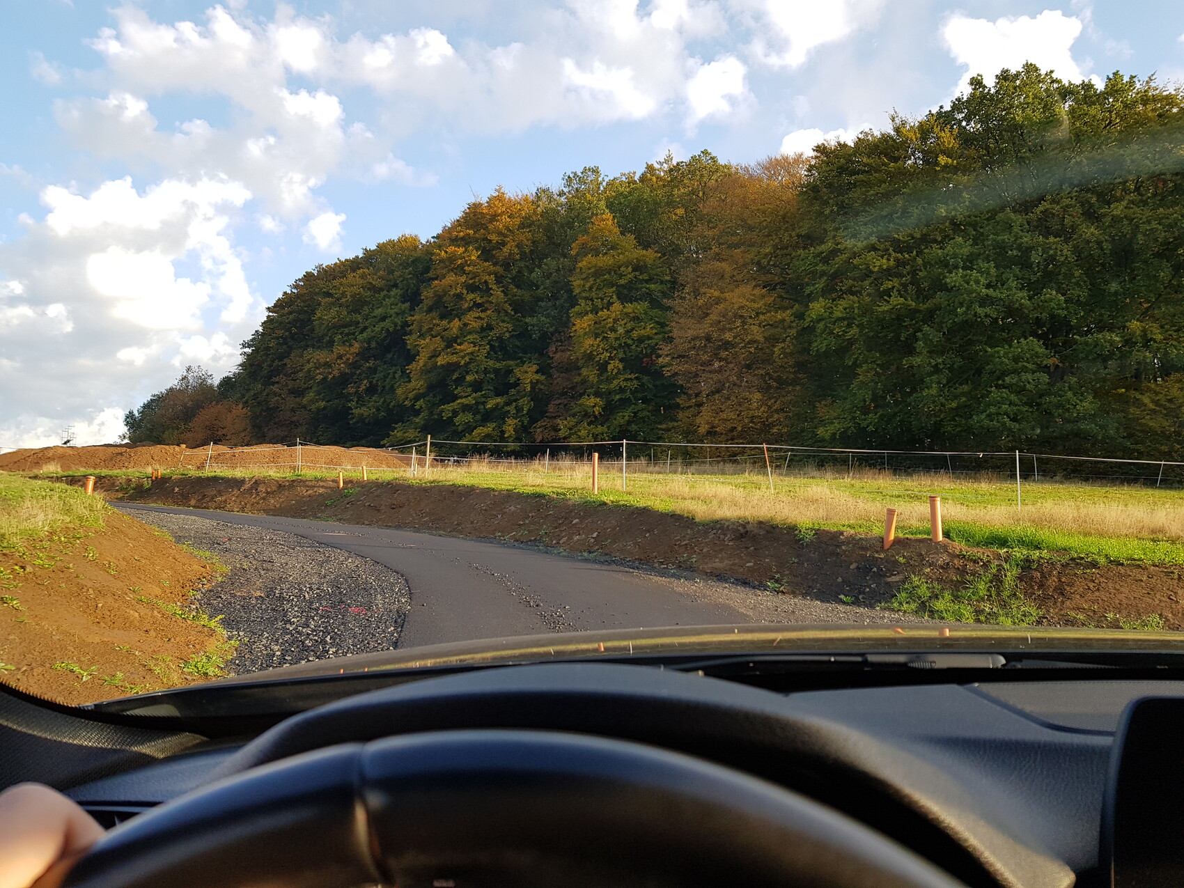 Fahrtaufnahme aus Auto: kurvige Baustellenstraße mit Kies, Gräben, Pfosten, Wald im Hintergrund.
