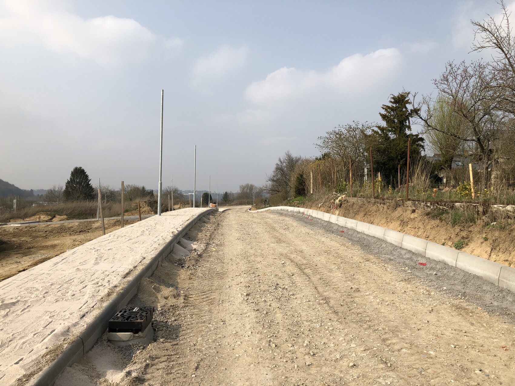 Unbefestigte Straßenbaustelle: Kiesweg mit Randsteinen, Pfostenreihen entlang, Bäume im Hintergrund.