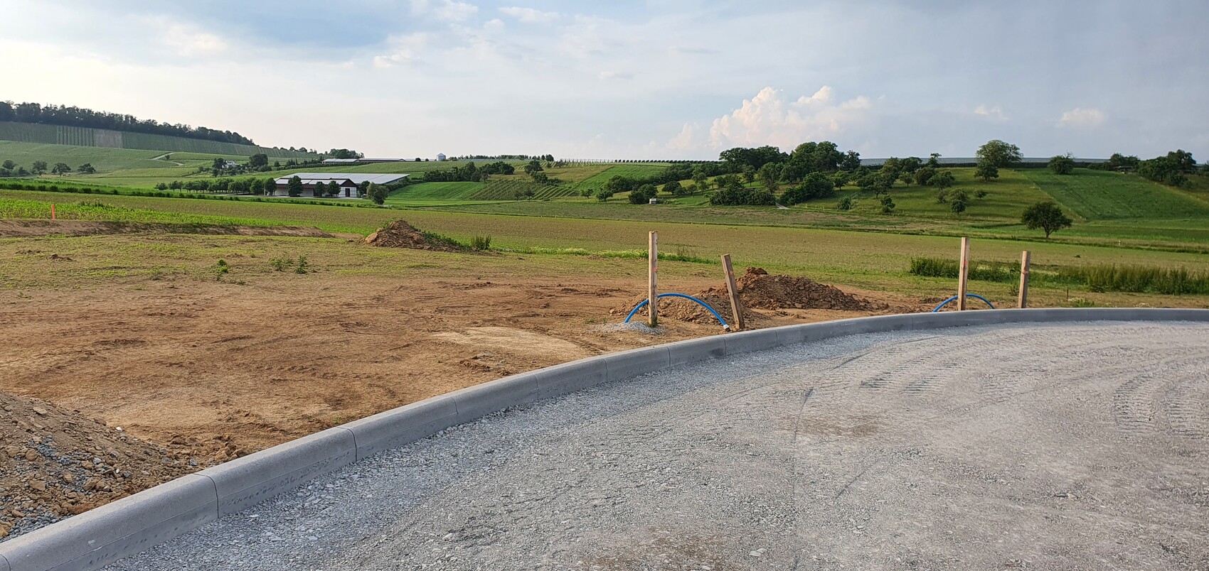 Kurvige Baufläche am Straßenrand, Kiesweg mit Bordstein und Baugrube; Felder und Hügel dahinter.