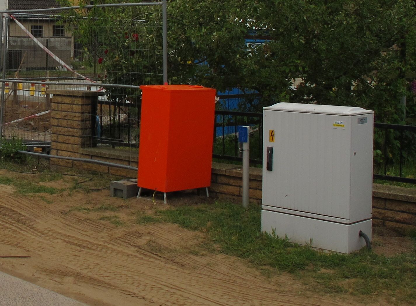 Orange Metallkiste neben weißem Elektroverteiler auf einer Baustelle mit Zäunen.