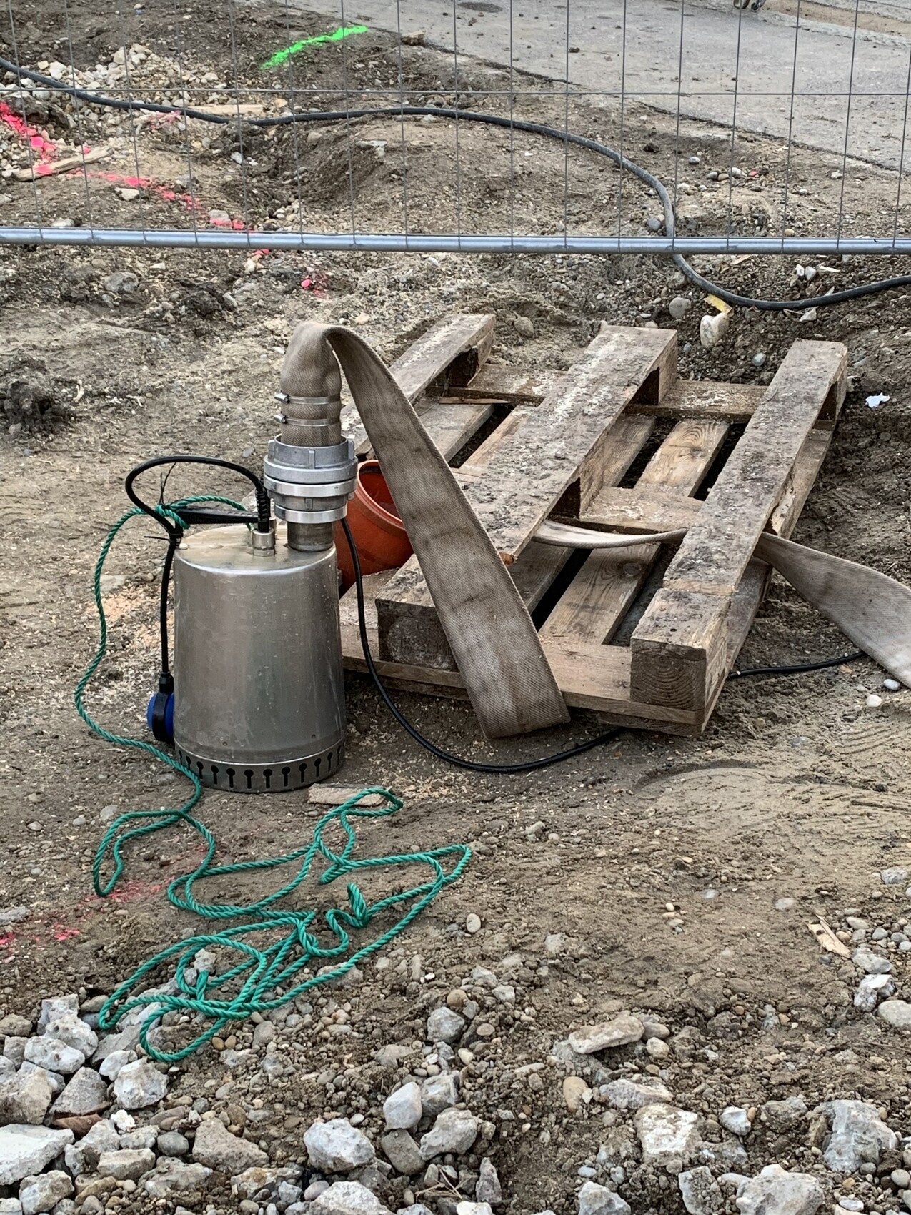 Baustelle: Tauchpumpe mit Schlauch neben einer Holzpalette auf Erde und Kies.