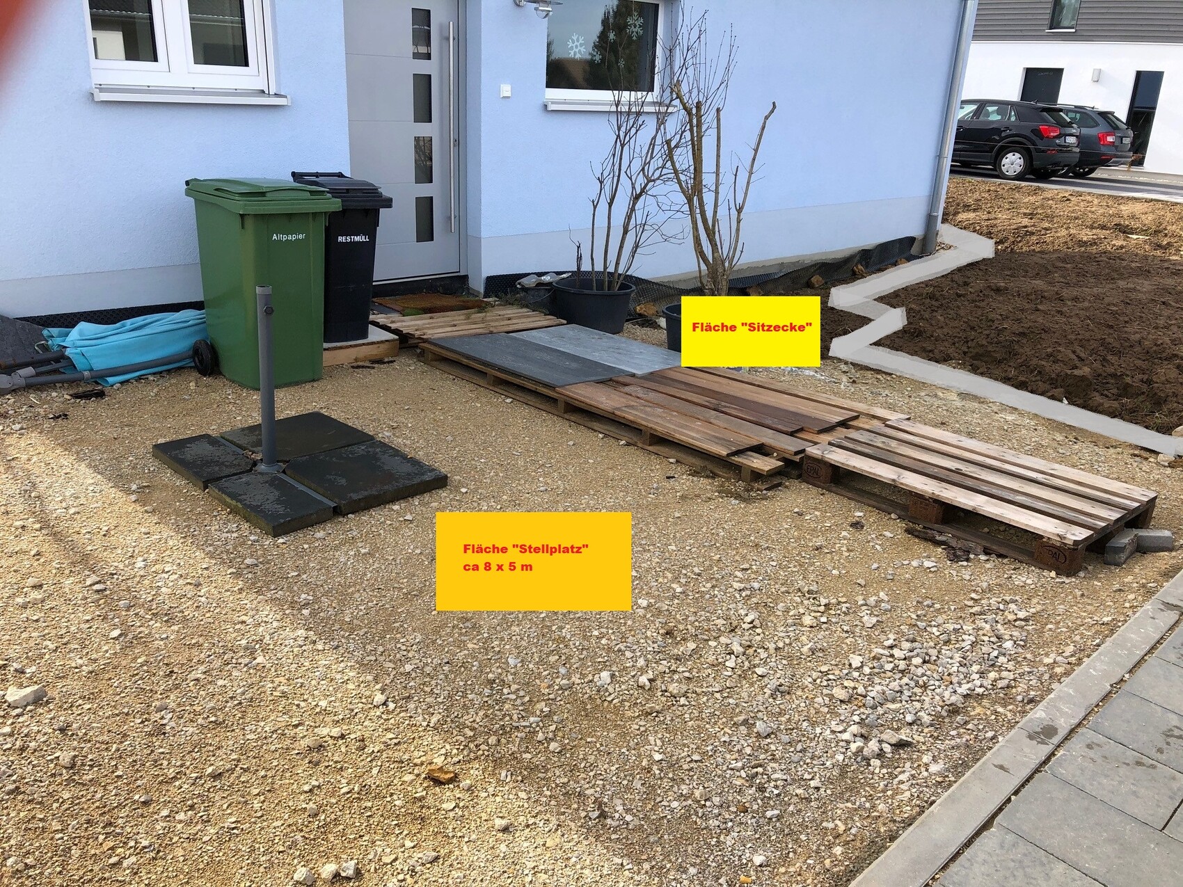 Außenbereich vor Haus mit Paletten, Holzplanken, Mülltonnen; Schild 'Stellplatz' & 'Sitzecke'.