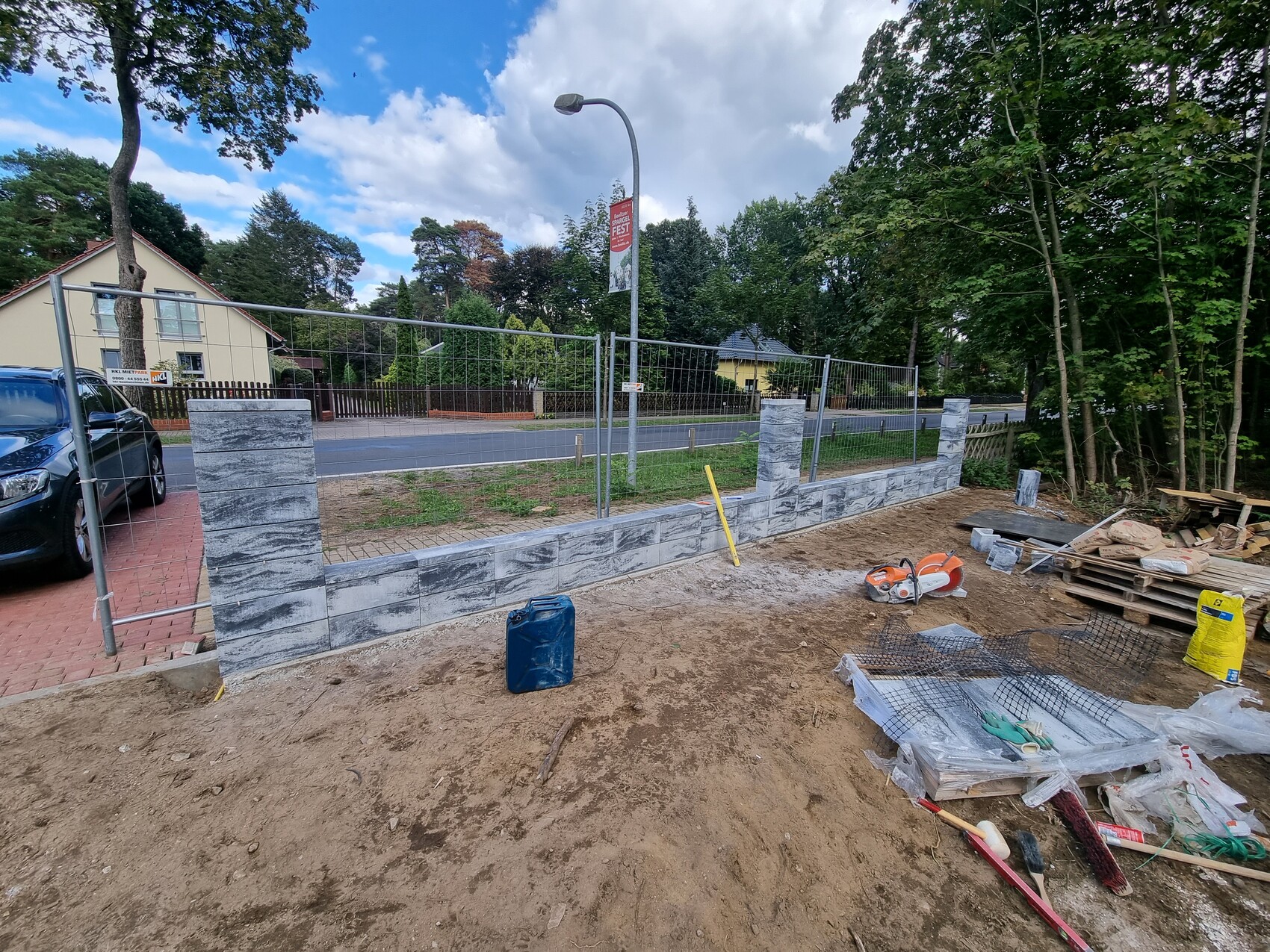 Baustelle: niedrige Steinmauer mit Metallzaun, Bauwerkzeuge, Paletten auf Sand; Wohngebiet dahinter.