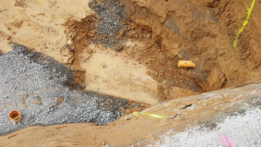 Baugrube mit Schlammwasser, orange PVC-Rohr, Kies und Erde; gelbes Sicherheitsband sichtbar