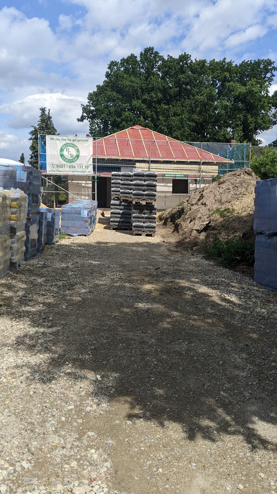 Baustelle mit gestapelten Paletten, Rohbau mit rotem Dach, Erdehaufen im Vordergrund.