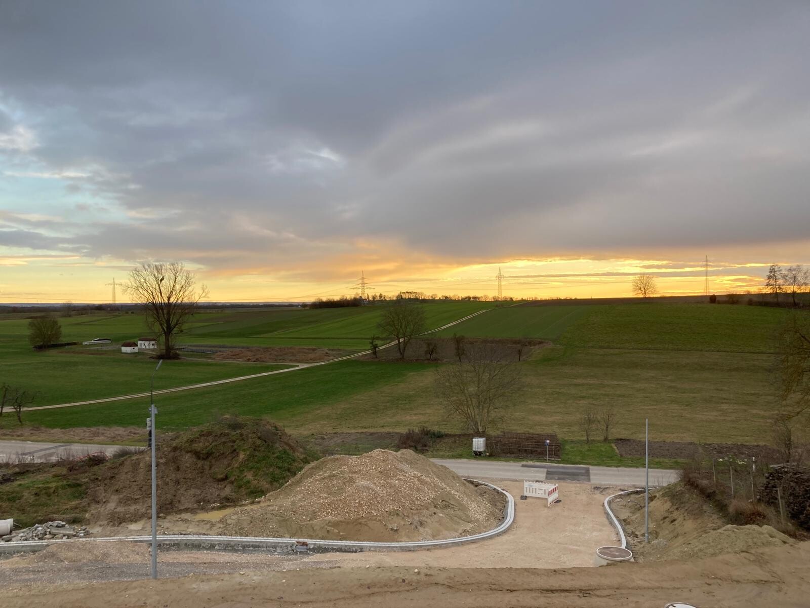Landschaft mit Feldern und Baustelle im Vordergrund, Sandhügel und Sonnenuntergang am Horizont