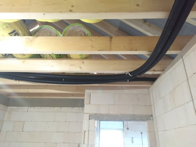 Baustelle mit Holzbalken-Decke, Dämmstoffrollen und schwarzen Kabelrohren entlang der Decke.