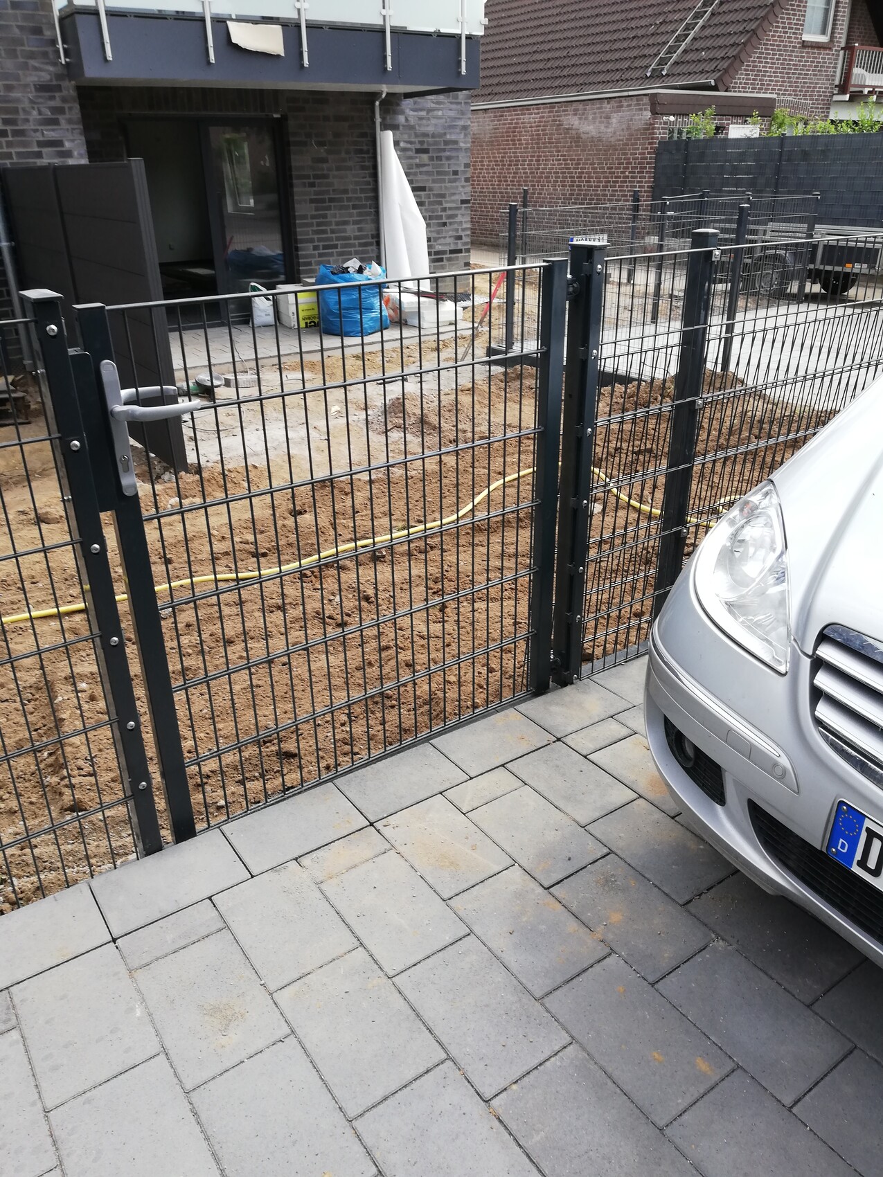 Baustelle hinter Metallzaun: Erde, Bauutensilien und blaue Müllsäcke; rechts parkt ein Auto.