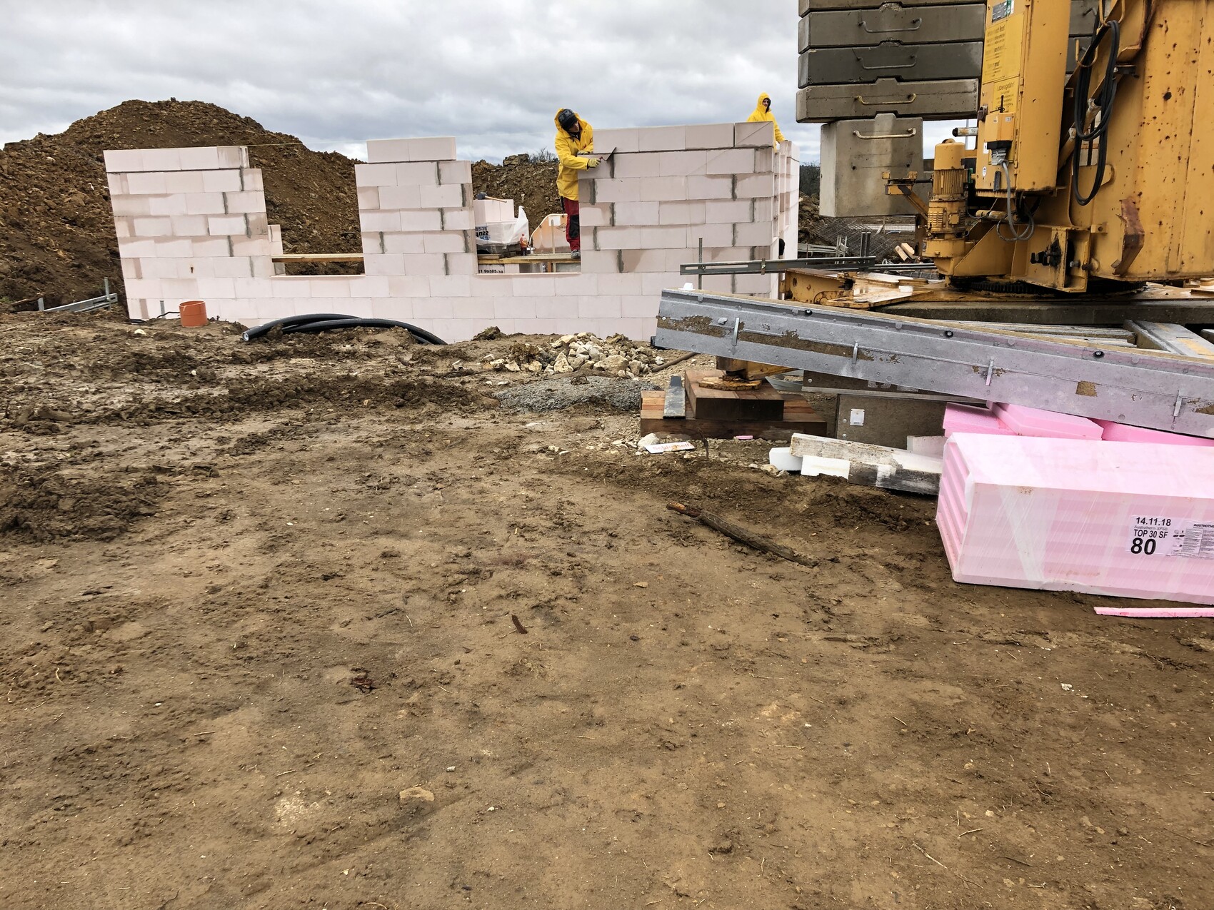 Baustelle mit zwei Arbeitern in Gelb an einer weißen Porenbetonwand; rechts Großgerät und pinke Dämmplatten.