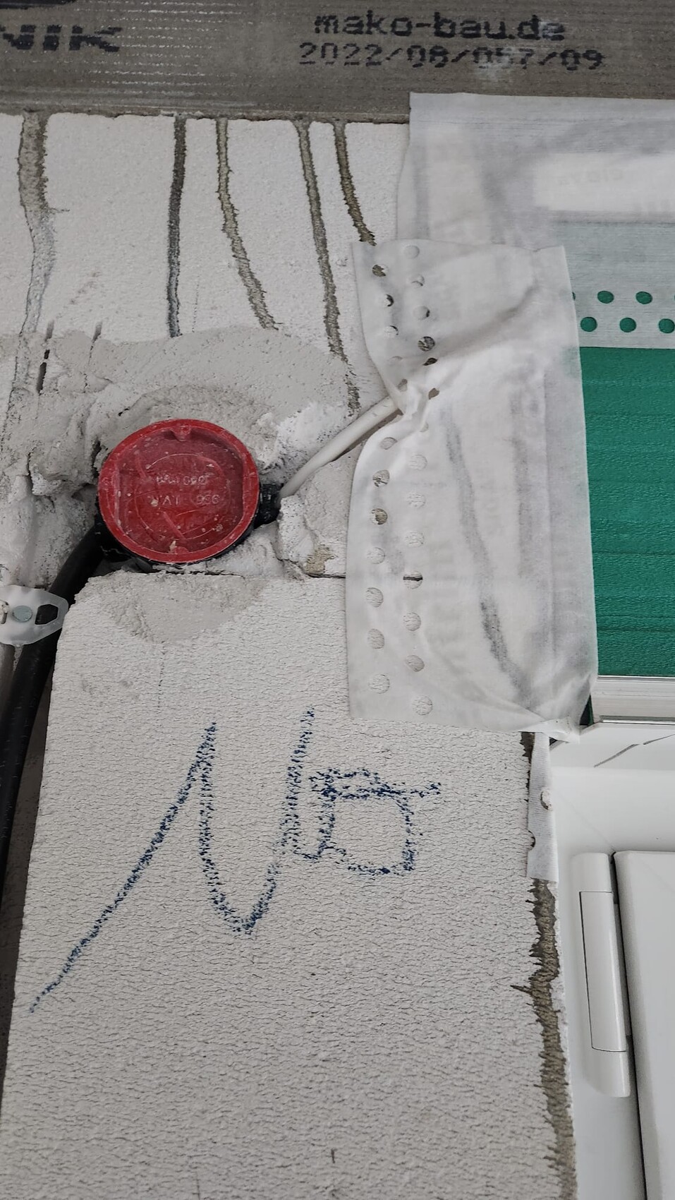 Nahaufnahme einer Bauwand mit roter runder Verschlusskappe am Rohr, Putzstaub und Markierungen.