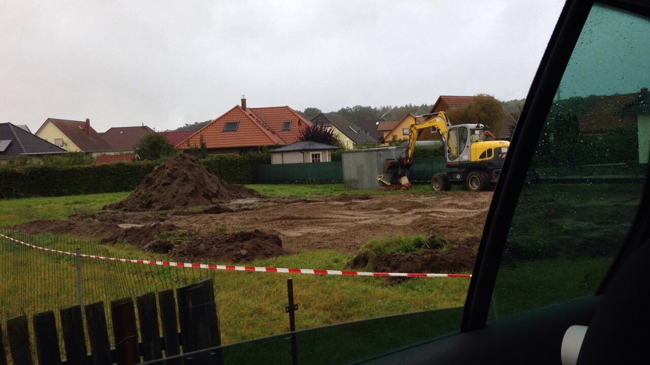 Baustelle mit Bagger, Erdhügeln und Absperrband; Wohnhäuser im Hintergrund.