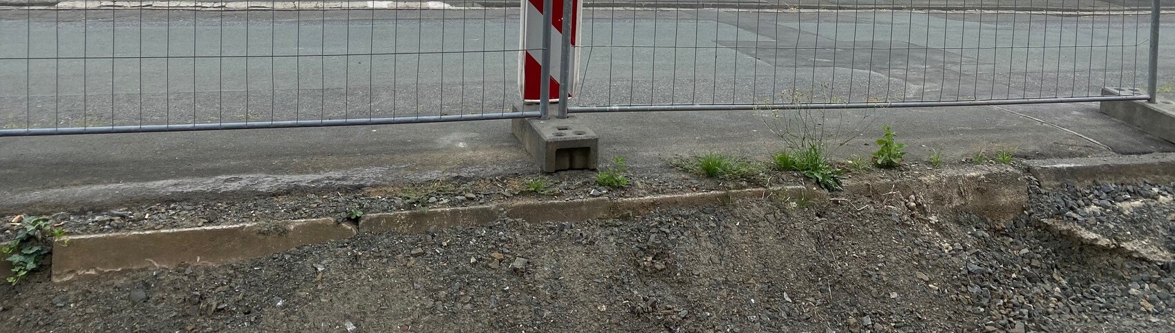 Metallzaun mit Betonfundament am Straßenrand; Gehweg mit Kies und Unkraut.