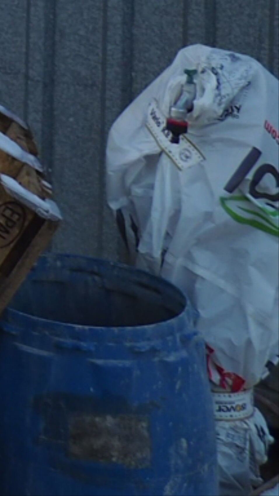 Blauer Müllcontainer neben einem weißen, zugebundenen Müllsack vor einer Wand.