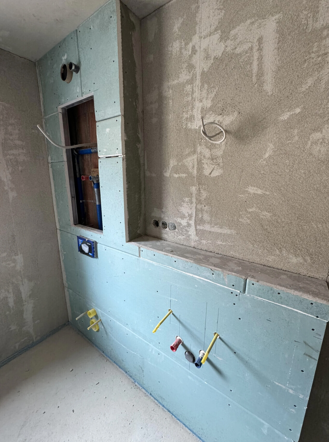 Unterbau eines Badezimmers: freiliegende Sanitärrohre hinter blauem Trockenbau, Wandnische sichtbar.