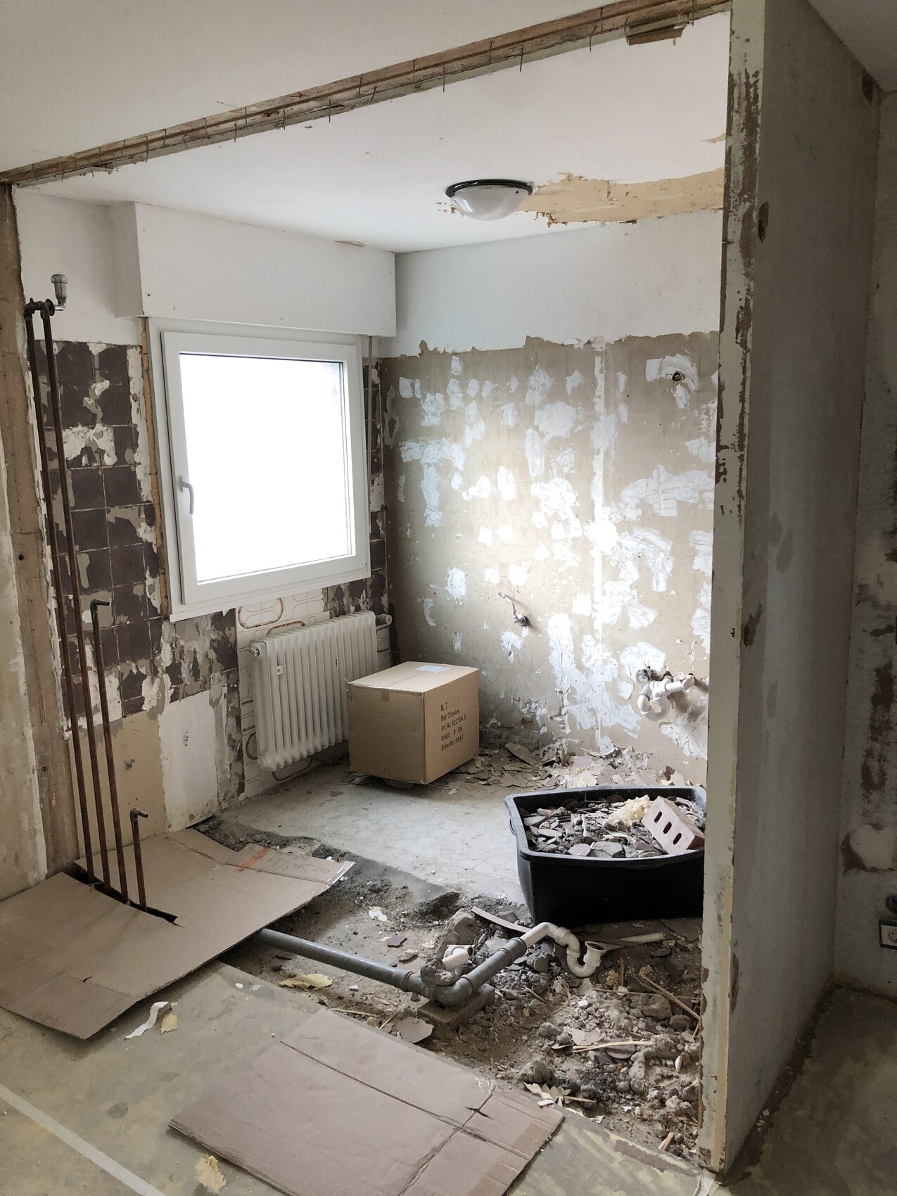 Renovierungsraum mit freigelegten Wänden, Fenster über dem Radiator, Kartonbox und Müllbehälter.