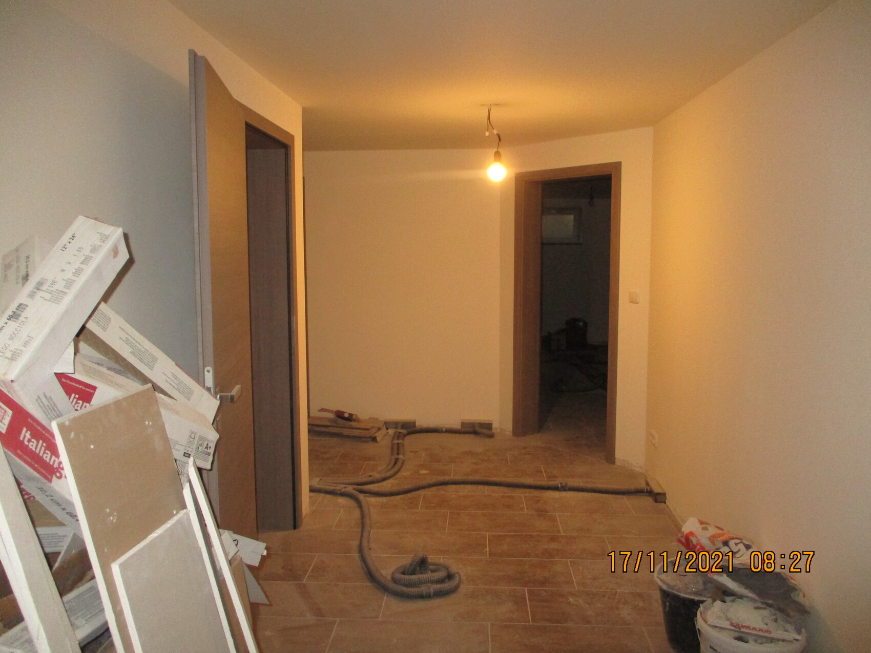 Heller Flur einer Baustelle: links Kartons/Trockenbau, lose Kabel, einzeln brennende Glühbirne.